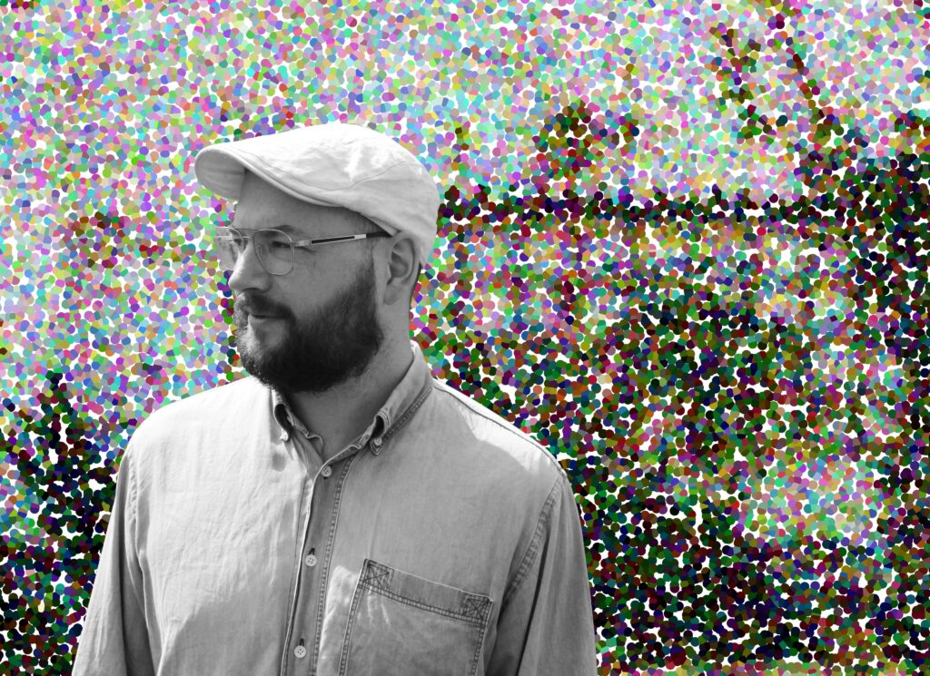 This is a black and white photo of Phil, wearing a flat cap and looking off to the left. He wears glasses and has a full beard. Behind him there is a digitally composed image of multi-coloured dots. 